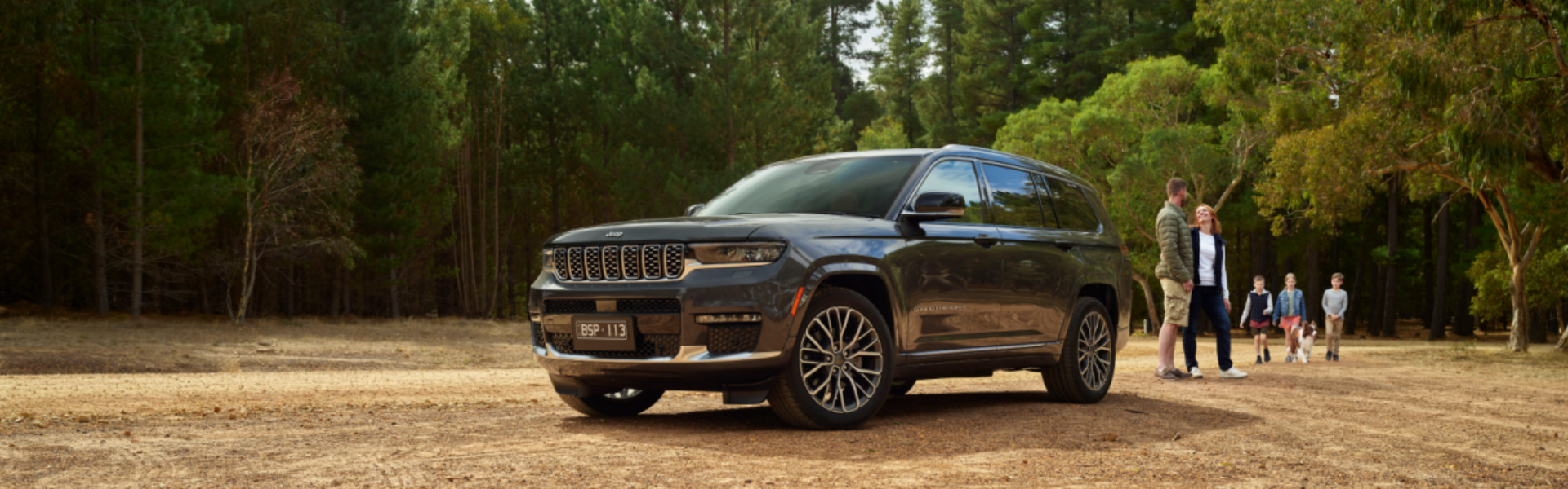 2024 Jeep Grand Cherokee parked in a scenic, wooded area with a family in the background enjoying an outdoor activity