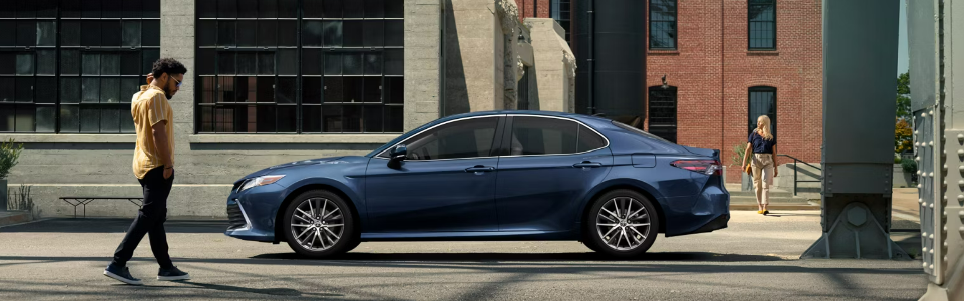 2024 Toyota Camry Hybrid positioned on a street in front of an industrial-style building with large windows FOR SALE