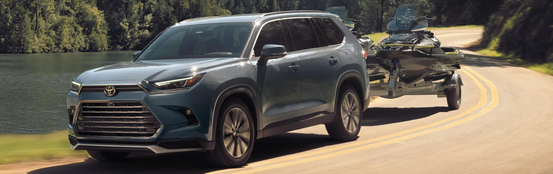 2024 Toyota Grand Highlander towing jet skis along a scenic lakeside road-Banner