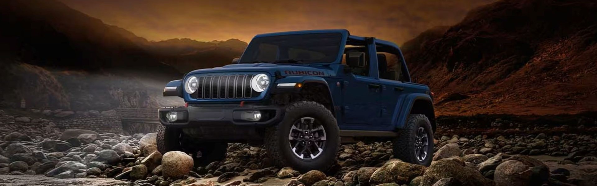 2026 Jeep Wrangler Rubicon X For Sale Banner - A blue 2026 Jeep Wrangler Rubicon X four-door, with its top removed and its headlamps on, parked on a rocky shoreline at the water's edge at night.